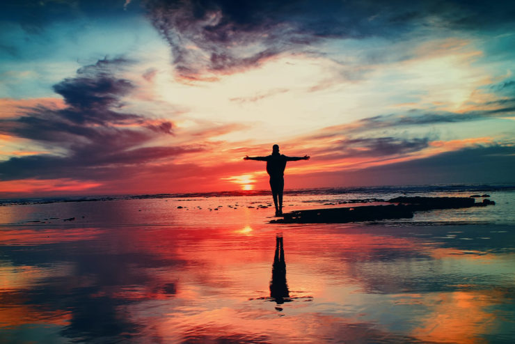 sunset over a body of water, with colorful clouds. The silhouette of a human figure stands near the center of the frame with their arms outstretched.